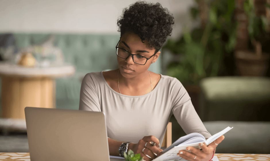 Woman with curly hair holding book and working on laptop